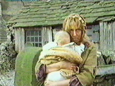 The woman stares out from the front of the barn, clutching her baby close
